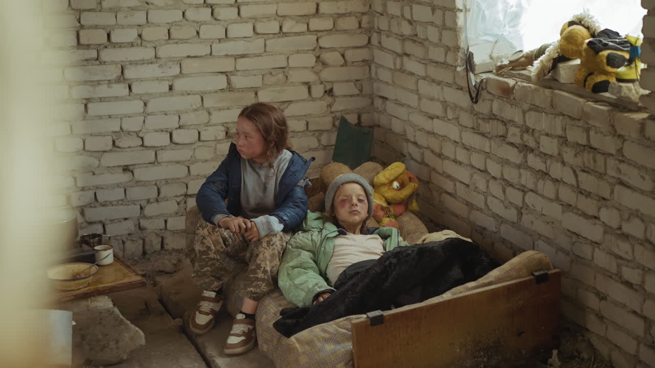 Two castaway children inside abandoned shelter, one sick lying on old mattress while other sits nearby staring away in confusion, faces marked with dirt, symbolising vulnerability of survival