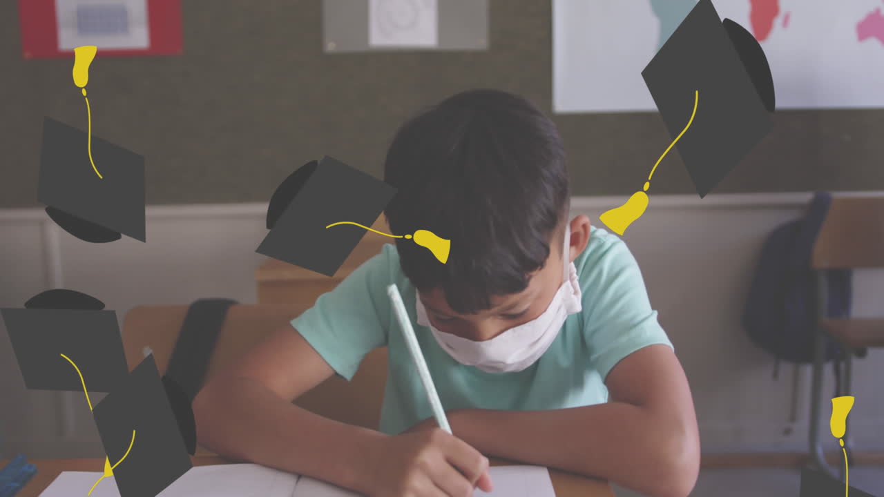 Animation of falling academic cap over boy wearing face mask sitting in classroom writing