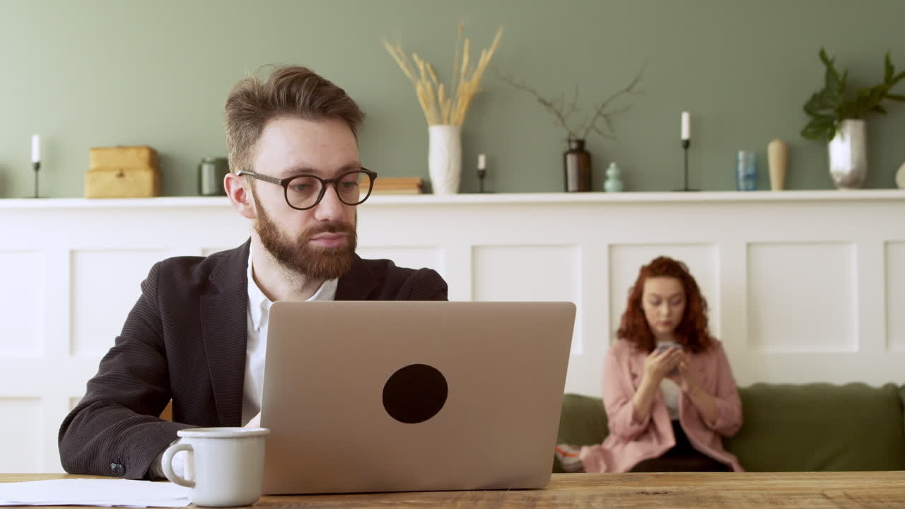 Bearded Man With Glasses Working On Laptop Computer While A Young Woman Using Mobile Phone Sitting On Sofa