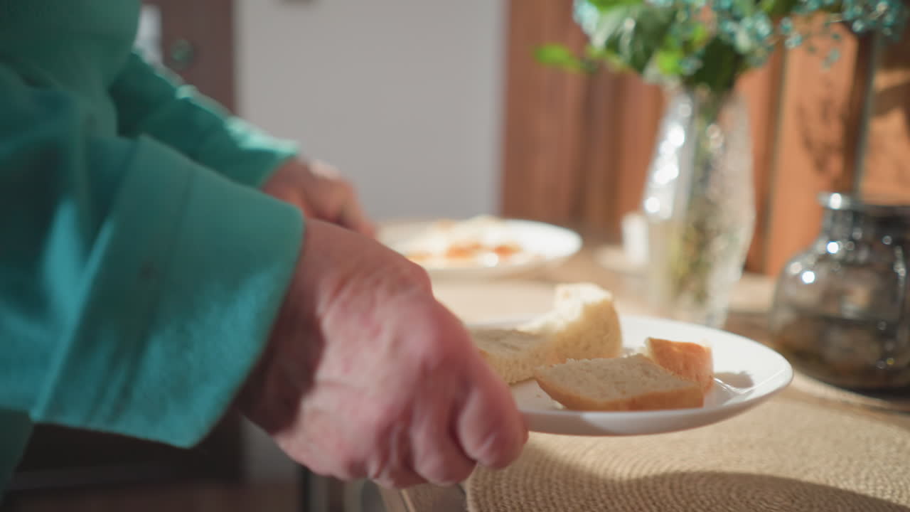 Elderly woman placing two plates with freshly sliced bread on dining table, preparing breakfast in cozy home kitchen. Interior with wooden walls, flowers, and placemats. Morning meal routine