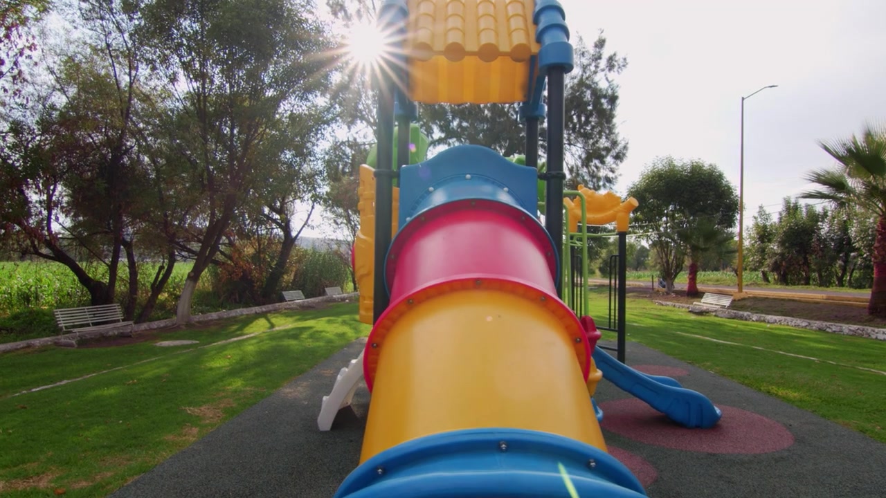 Vista ravvicinata di uno scivolo a tubo colorato in un parco giochi, situato in un soleggiato parco pubblico, con erba, alberi, panchine e un vivace scenario di svago familiare.