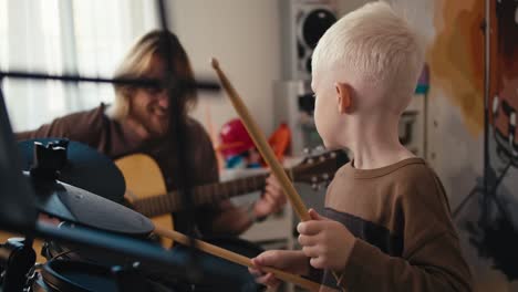 toma de primer plano de un feliz niño albino con cabello blanco y un corte de pelo corto tocando batería electrónica usando palos especiales para instrumentos de percusión y su padre tocando una guitarra acústica con él, un feliz hombre rubio con barba y gafas en la habitación