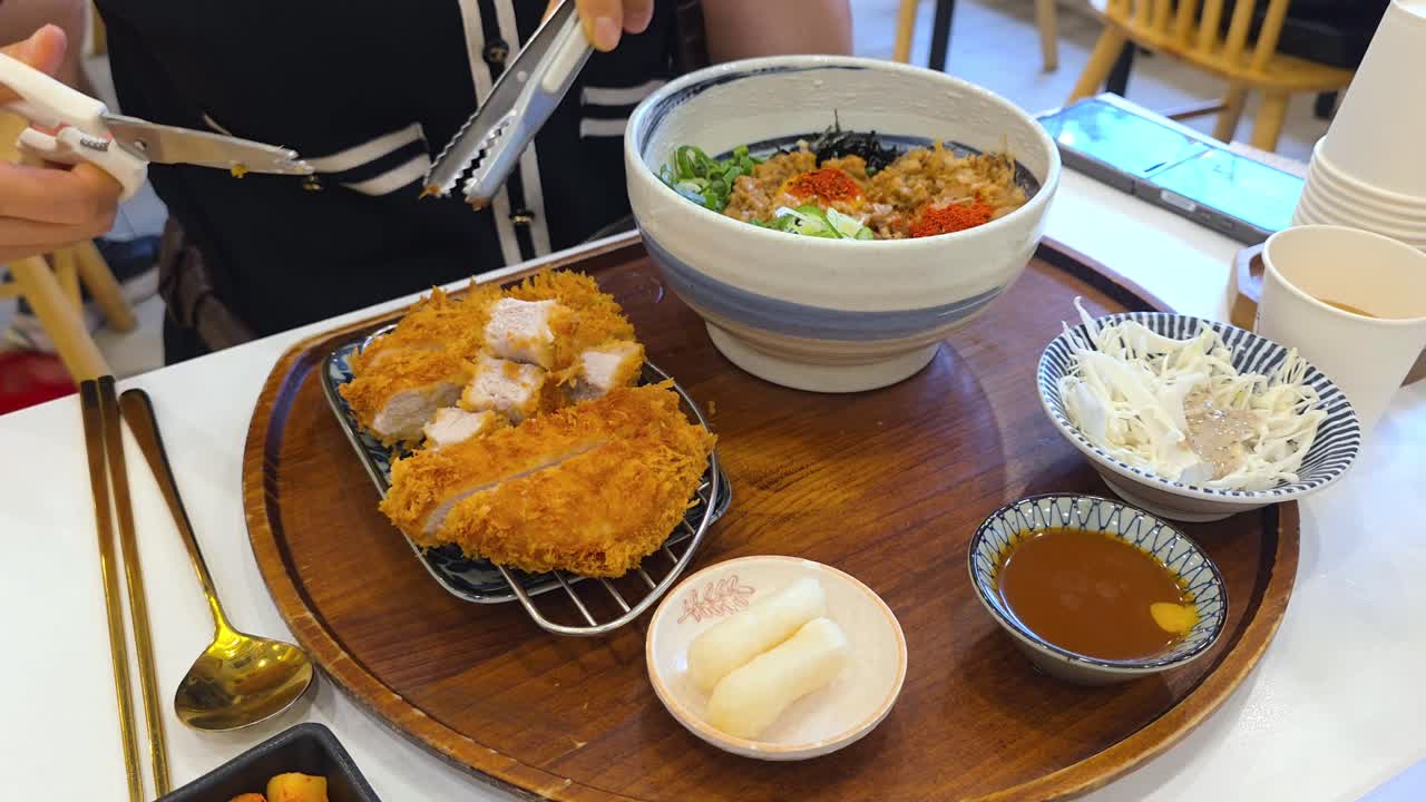 A person uses scissors to cut a crispy pork cutlet served with rice, shredded cabbage, sauce, and pickled radish in a modern Korean restaurant setting