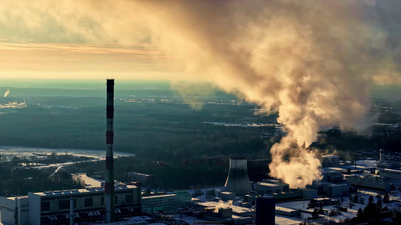 Ripresa aerea di una centrale elettrica e termica invernale all'alba, con un alto fumaiolo e una torre di raffreddamento che rilasciano una colonna di vapore sopra i tetti innevati e la foresta ripresa da un drone