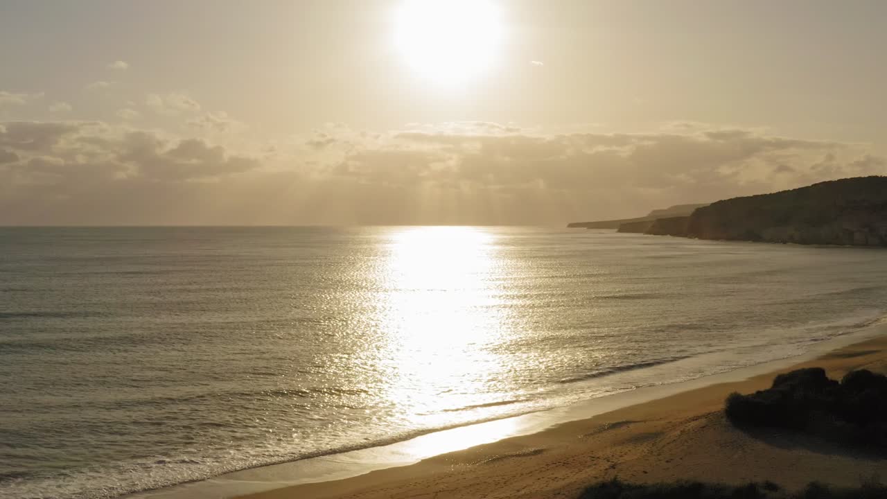 Plano aéreo cinematográfico de pôr do sol sobre o oceano. Drone recuando para revelar a praia.