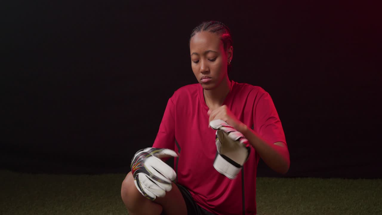 Donna afroamericana inginocchiata sull'erba sintetica con una maglietta rossa che allaccia i guanti da portiere prima dell'allenamento.