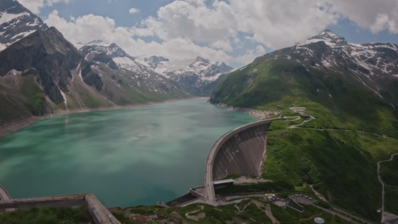 A nice viewpoint overlooking a turquoise reservoir surrounded by majestic, snow-capped mountains. A rustic wooden fence and rugged foreground rocks enhance the natural charm of this alpine landscape.