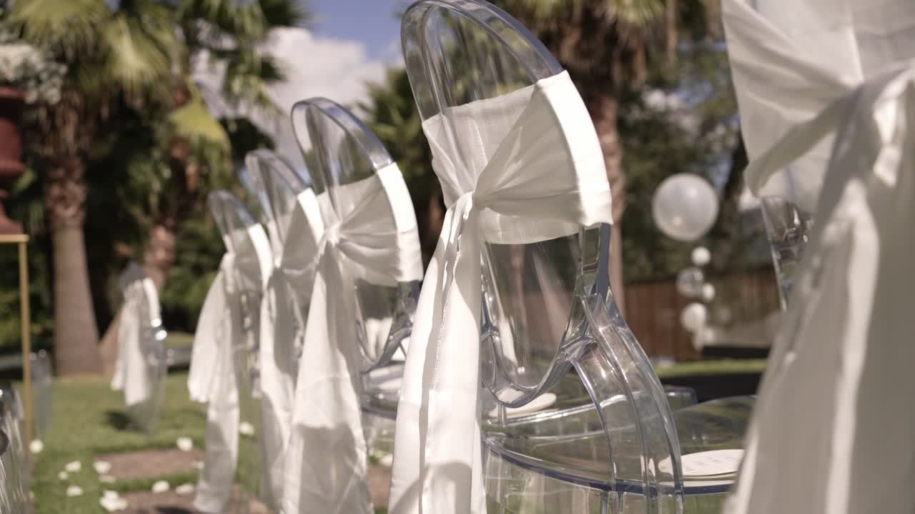Transparent wedding chairs with white sashes in outdoor setup under sunny skies