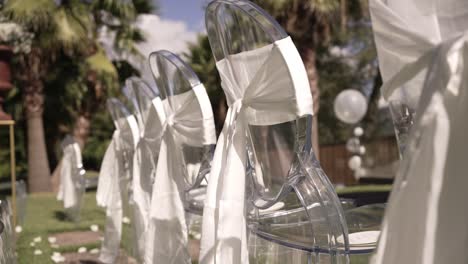 transparent wedding chairs with white sashes in outdoor setup under sunny skies