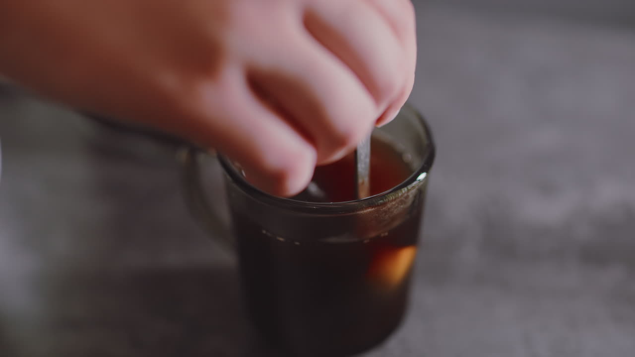 Close up of lady hand stirring coffee after adding sugar into transparent cup, showing swirling motion of spoon mixing drink on table surface during beverage preparation routine indoors