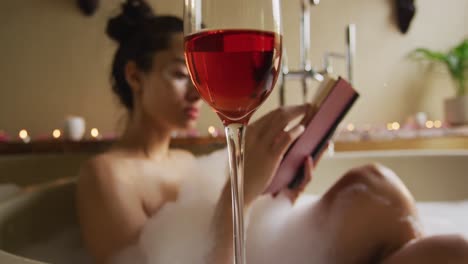 Copa-De-Vino,-Con-Una-Feliz-Mujer-Birracial-Con-Vitíligo-Leyendo-En-Un-Baño-Con-Espuma-En-El-Fondo