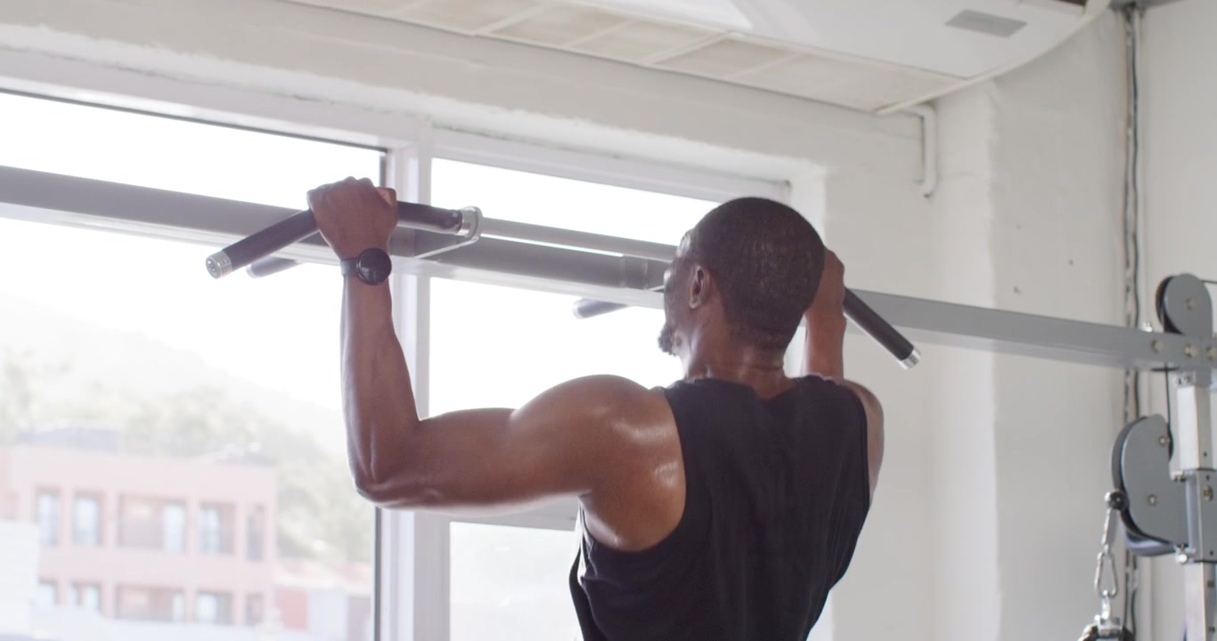 African American man wearing black tank gripping pull-up bar, doing pull-ups at gym for strength