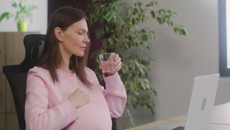 pregnant woman caressing her belly and drinking water while working in the office