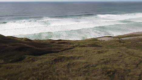 un dron de vuelo bajo de hermosas dunas de cornualles que se dirigen hacia el mar con impresionantes olas rodando hacia una playa de arena dorada