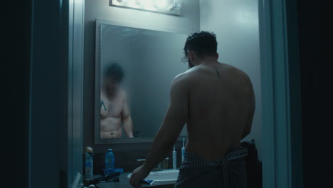 man walks out towards mirror, towel wrapped around, wipes steam off and views his reflection in the mirror