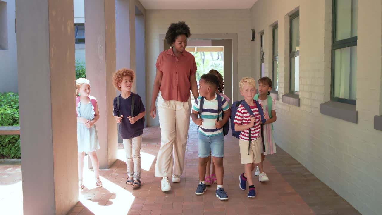 African American woman from doorway leading six children adjusting backpacks while holding tablet