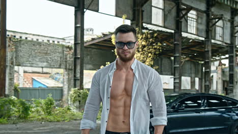 handsome man with a pistol in ruined place. young bearded man in glasses holds the handgun and looks at camera on luxury car background.
