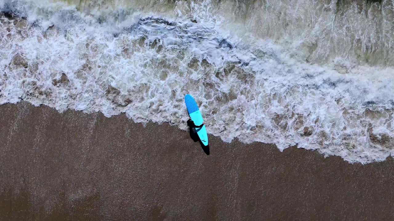 una vista de arriba hacia abajo de un surfista caminando fuera de su tabla de surf en el océano en un día soleado
