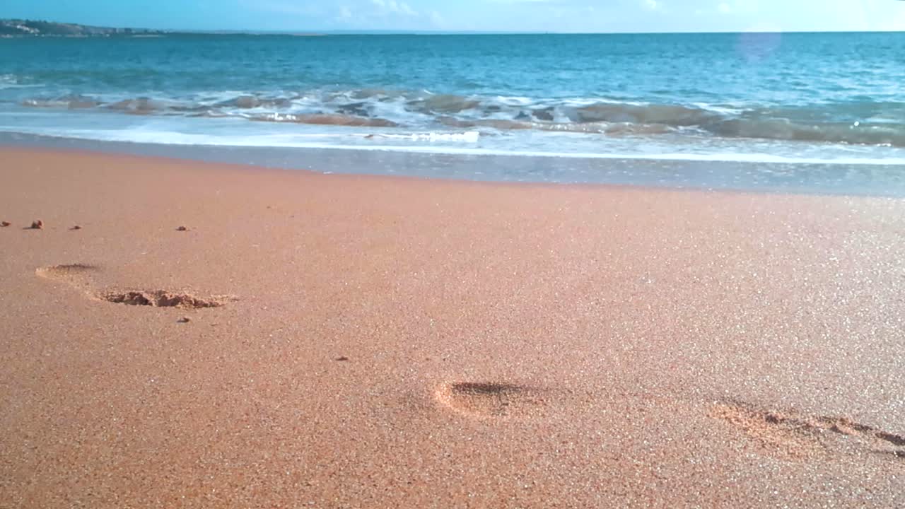 Vista dal basso di impronte nella sabbia morbida e umida con dolci onde atlantiche che si infrangono su una soleggiata costa portoghese