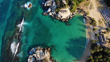 Aerial-view-above-shallow,-turquoise-sea,-a-beach-and-a-resort-on-the-coast-of-Tayrona-national-natural-park,-sunny-day,-in-Colombia---top-down,-drone-shot