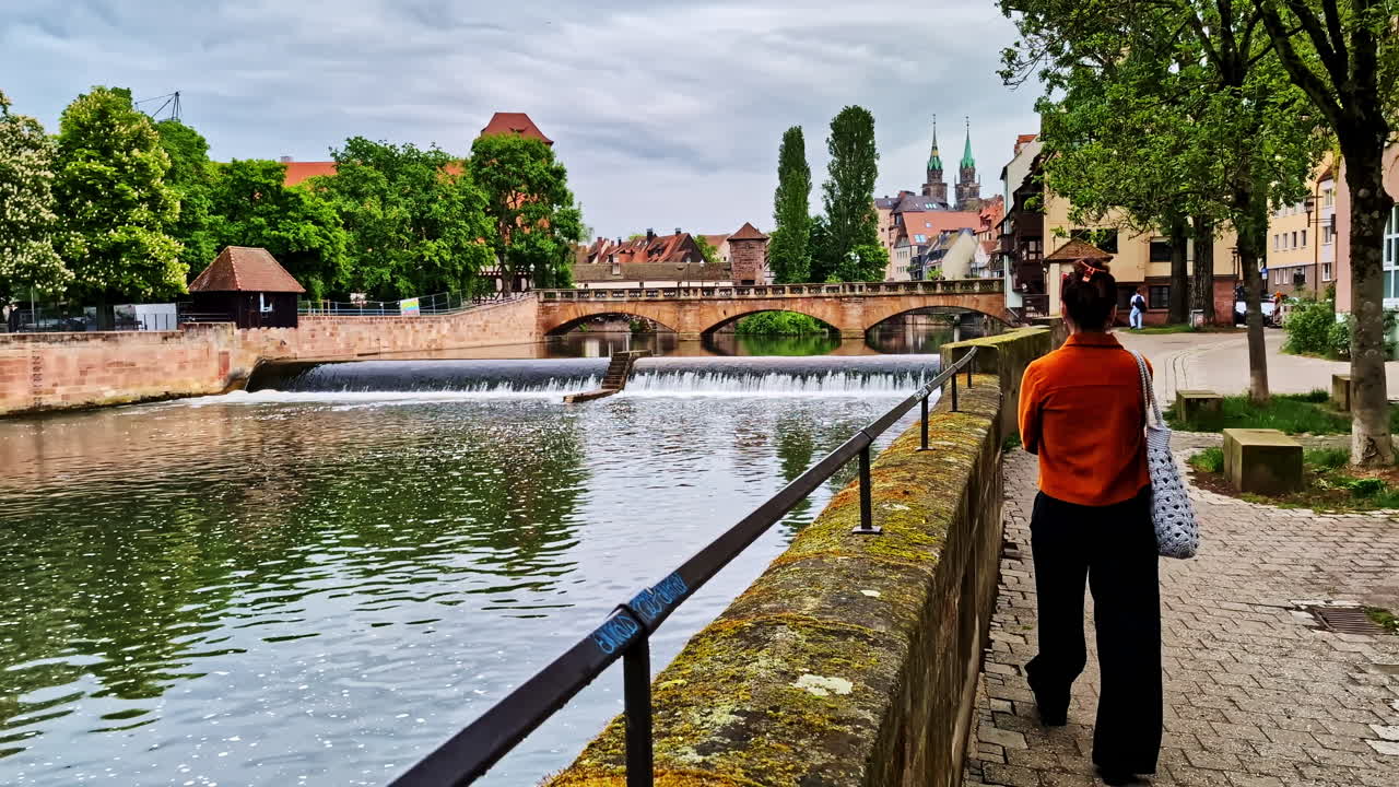 Woman Walking On The Riverside Path Looking To The Pegnitzwehr And Maxbrucke in Nuremberg, Germany. - wide shot