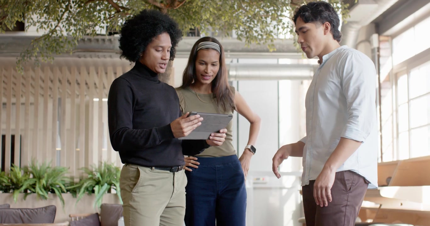 Diverse coworkers entering office lounge, left presenter presenting tablet and guiding work review