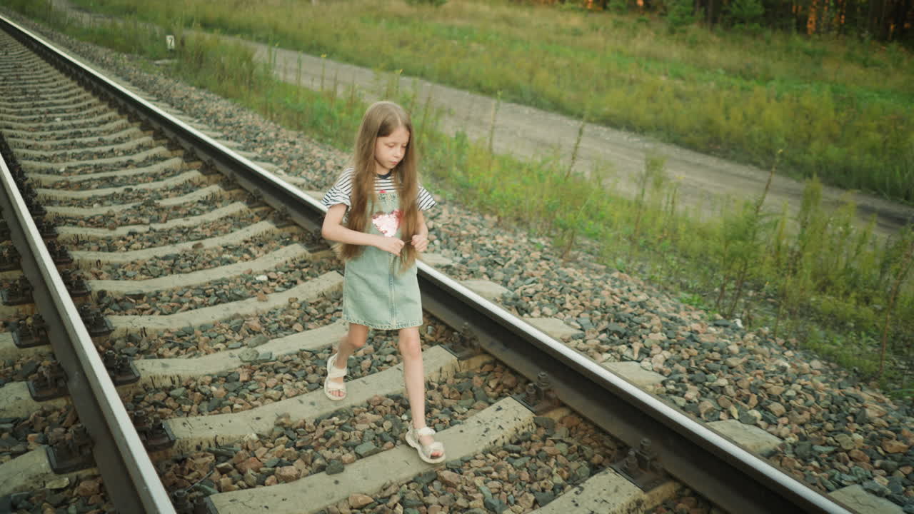 young girl with long hair walking thoughtfully along rail track holding grass strand, wearing striped top and denim dress, surrounded by green vegetation and rocky sleepers in rural outdoor setting