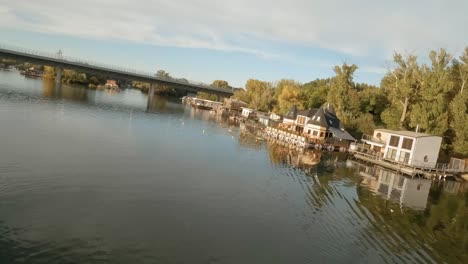 Drohnenaufnahmen-Von-Farbenfrohen-Hausbooten,-Die-An-Einem-Ruhigen-Fluss-Mit-Einer-Brücke-Im-Hintergrund-Festgemacht-Sind