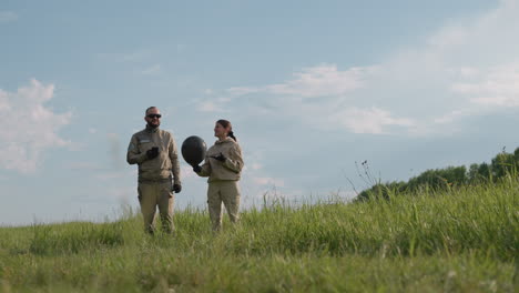 man and woman walking through tall grass field under blue sky wearing protective suits gloves, woman holding black balloon while they converse amid serene rural landscape