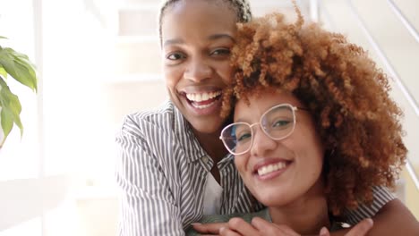 portrait of happy diverse lesbian female couple on stairs and embracing at home in slow motion