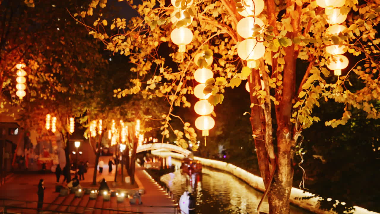 Cena noturna com iluminação acolhedora, lanternas penduradas em árvores junto a um canal