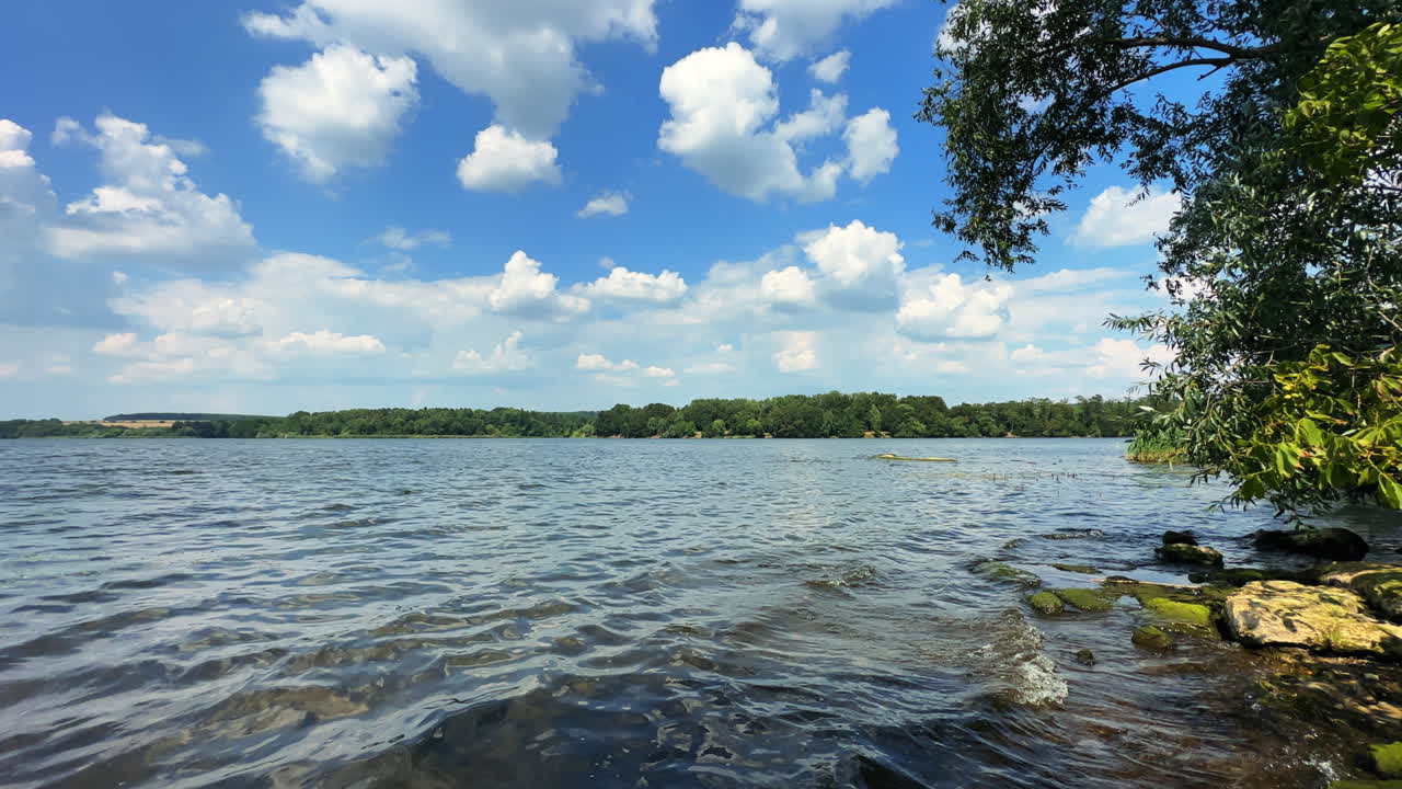 Little waves roll to the rocks on the river bank. Beautiful waterscape under blue sky with clouds on summer day.