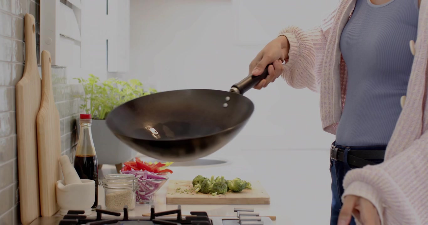 Mid adult woman reaching for oil bottle and pouring oil into wok while tossing vegetables