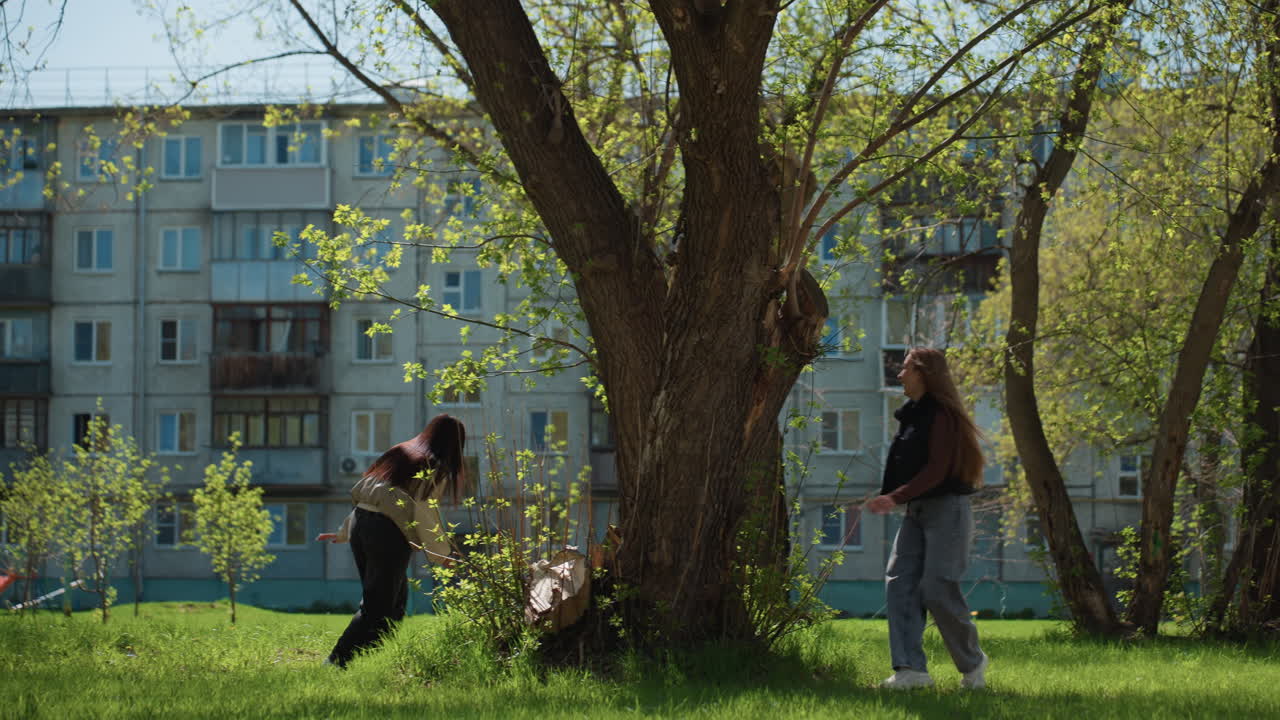 젊은 백인 여성과 아시아 여성 두 명이 햇빛이 비치는 도시 안뜰에서 나무 아래를 걷고 있어요. 배낭을 메고 천천히 재회하며 아파트 건물을 지나고, 푸른 잔디와 부드러운 바람이 불고, 조용한 대화가 오가는 평화로운 분위기입니다.