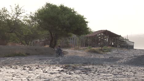 Man-and-passenger-arriving-to-a-wood-shack-in-Popoyo-beach-Nicaragua-riding-a-motorcycle-over-the-sand-with-legs-down-to-stabilize,-Pan-right-shot