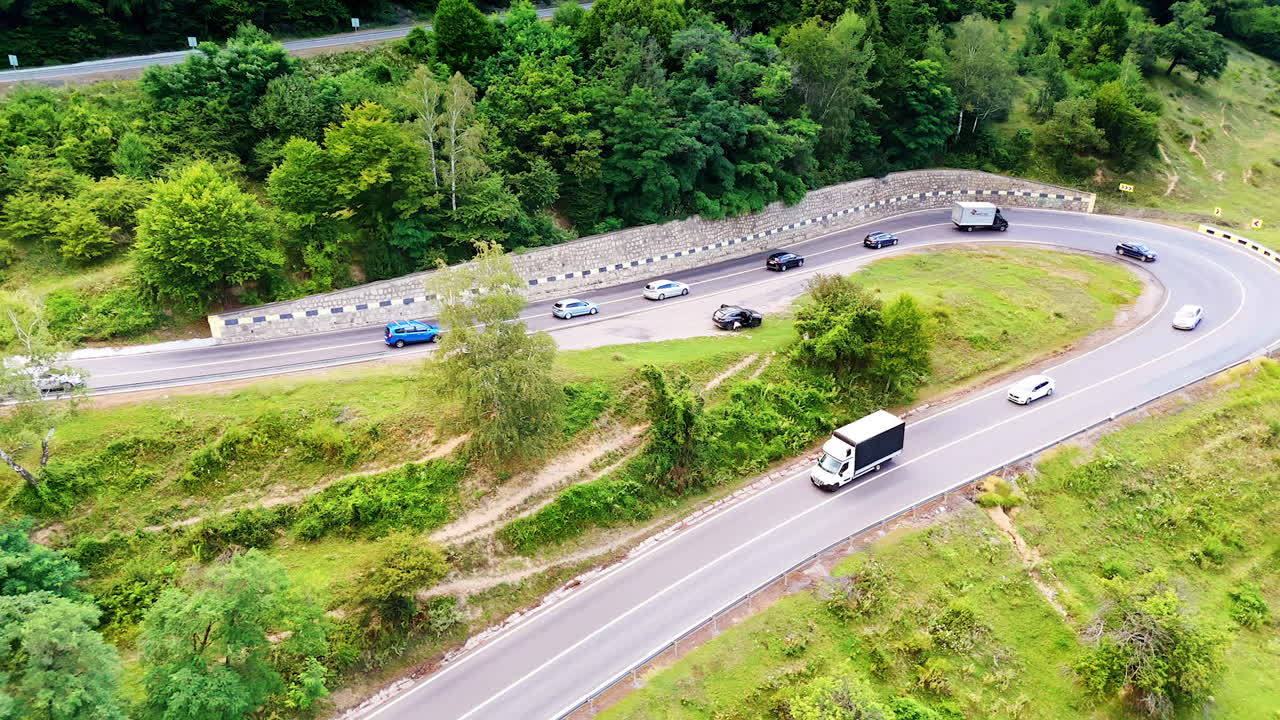 Traffic flows along a winding rural road. Various vehicles navigate a serpentine road surrounded by lush green vegetation on a sunny day