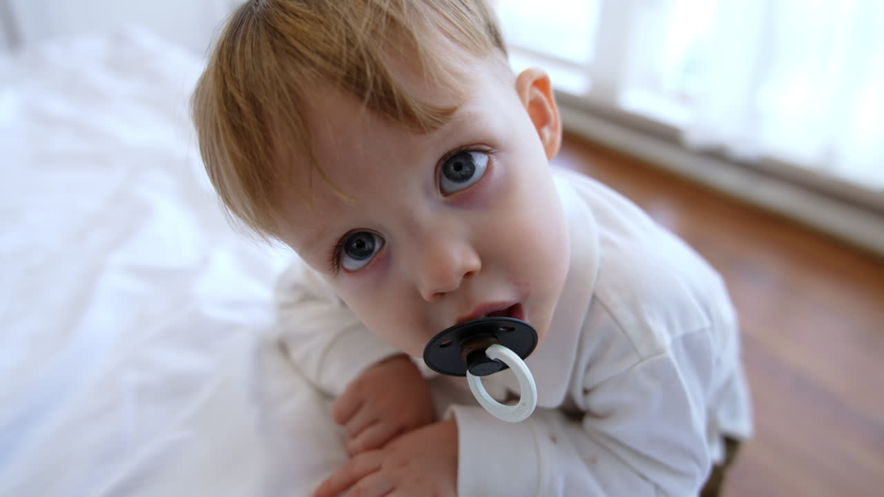 Adorable baby boy stands at the bed looking at camera. Cute big-eyed toddler sucking a pacifier. Close up.