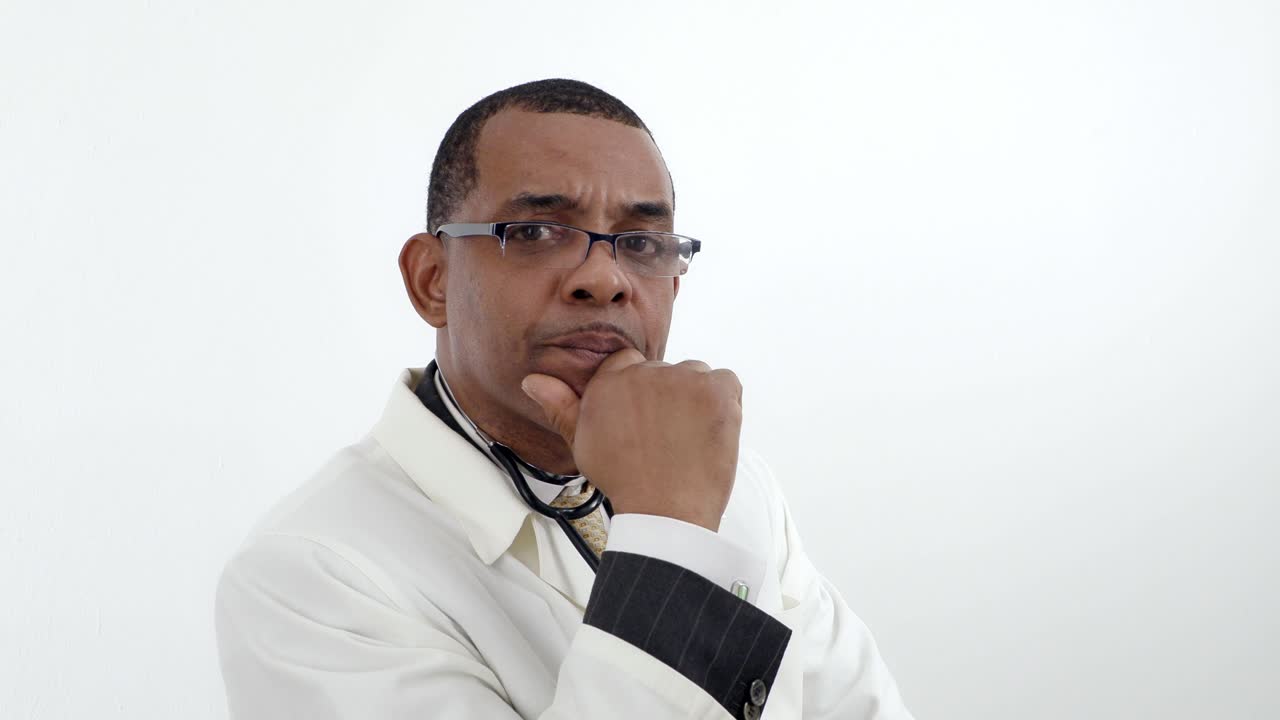 Jamaican African-American man doctor in white lab coat  in glasses, turning head looking at the camera with serious concern over white wall background isolated.
