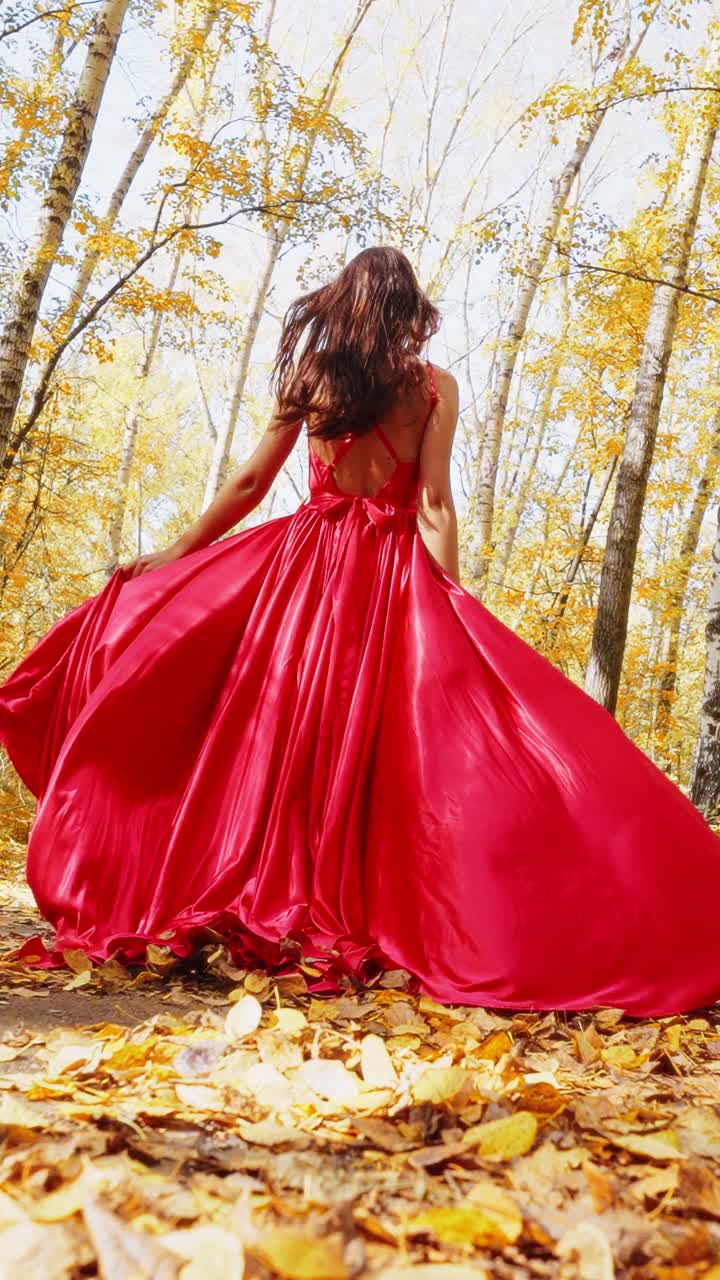 Elegant woman in a flowing red gown walks through a picturesque autumn forest, surrounded by golden leaves and vibrant trees under bright sunlight, creating a stunning natural backdrop