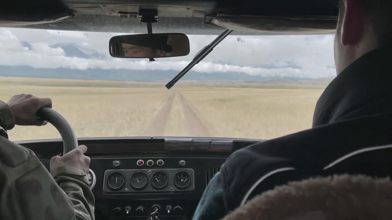 Punto di vista dall'interno di un veicolo che guida su una strada sterrata attraverso l'ampia Steppa di Kurai, Repubblica dell'Altai, Russia, con montagne distanti e cielo nuvoloso