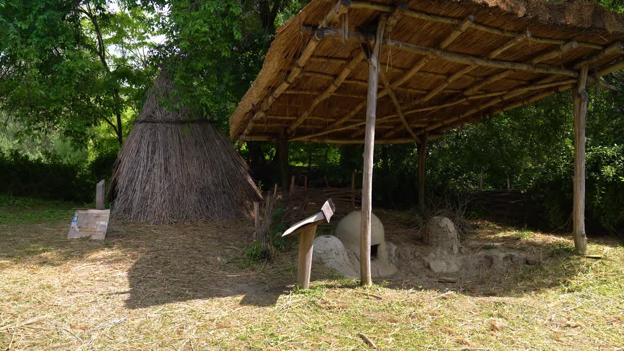 Thatched roof shelter and primitive clay oven near straw hut in open-air museum setting