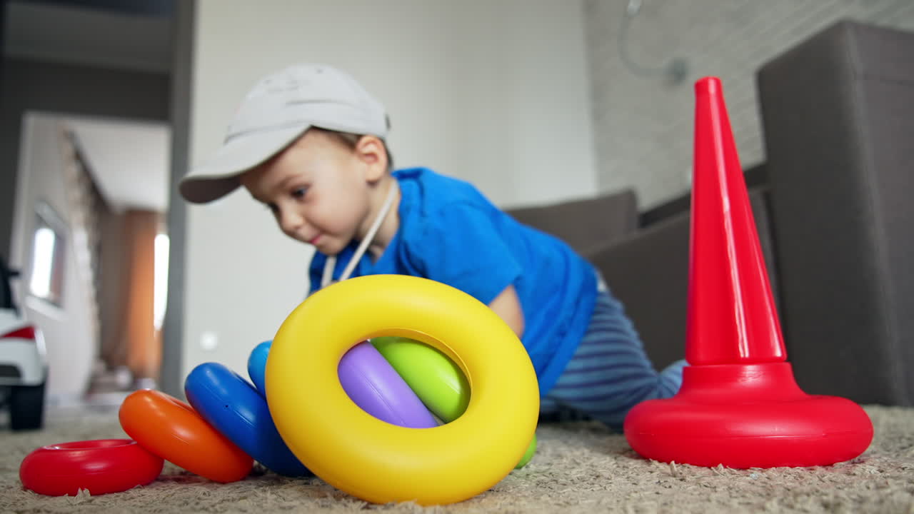 Funny little kid in cap crawls around the toy pyramid. Toddler stands up and goes to another toy. Low angle view.