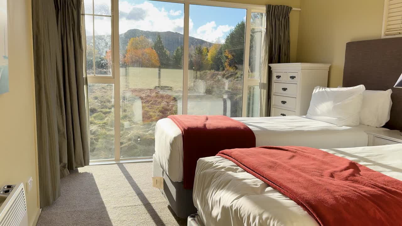 Camera moves through an opening door into a bright, modern bedroom with large windows, revealing a picturesque lakeside and mountain landscape outside