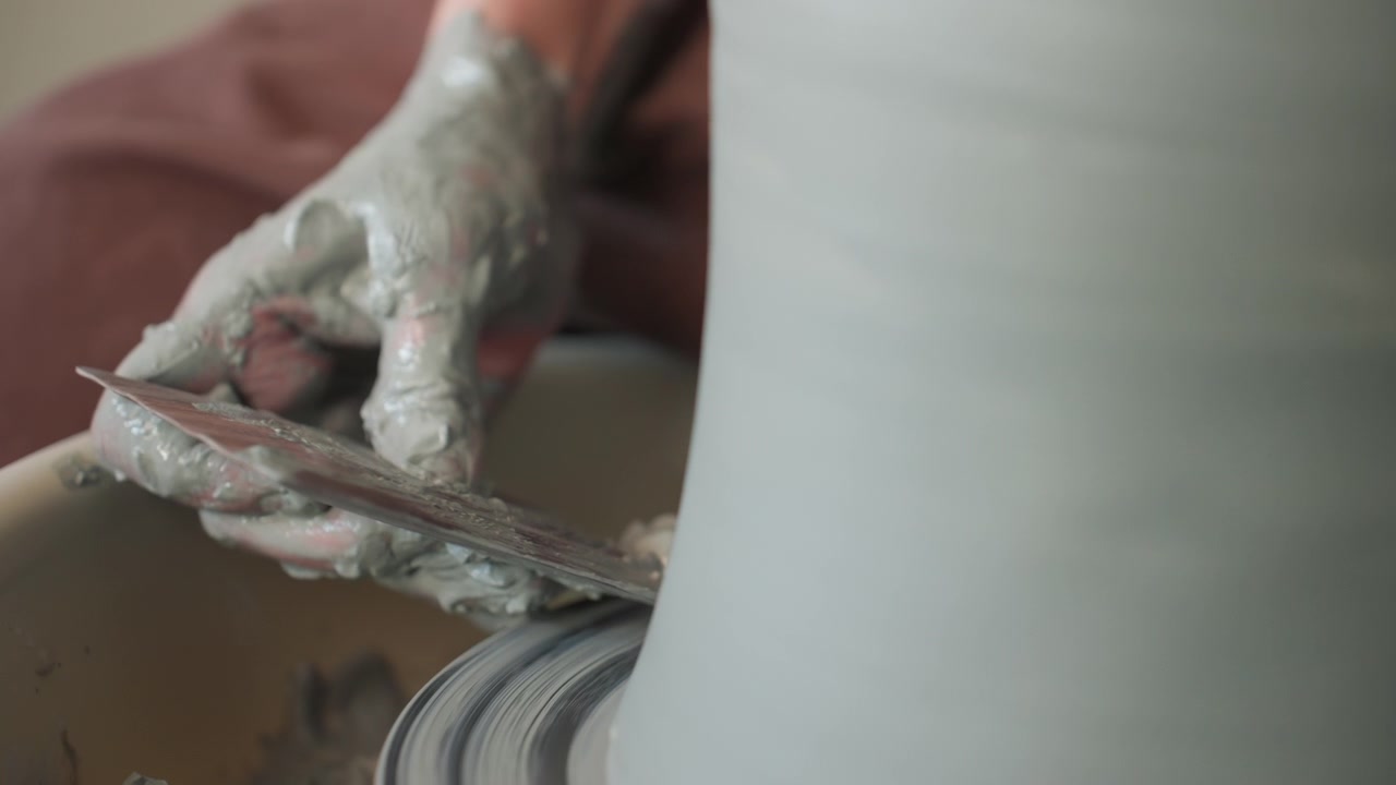 Pottery workshop caucasian finisher trimming clay base with wire tool while wheel rotates, steady hands cutting foot ring, dust flecks and careful precision, quiet finishing moment in studio