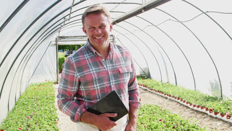 portrait of mature male owner of garden center holding digital tablet in greenhouse