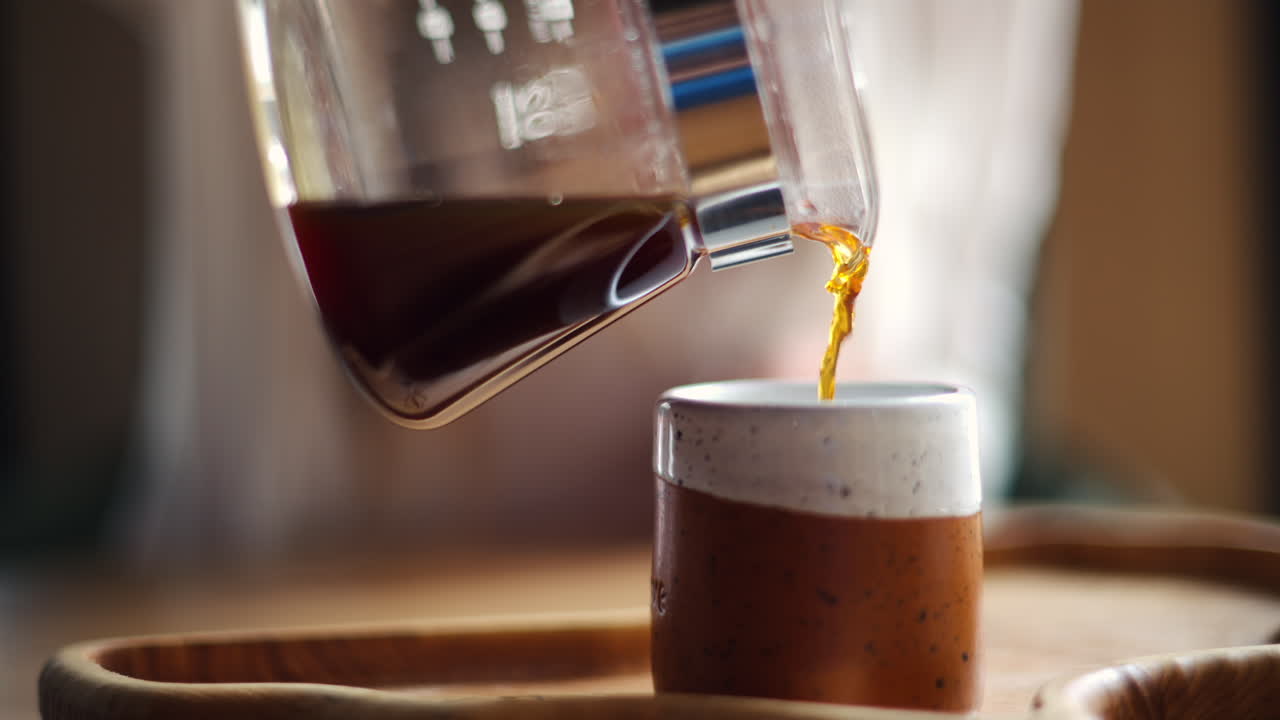 Close up of a woman pouring coffee in a cup from a glass pot standing on a wooden tray
