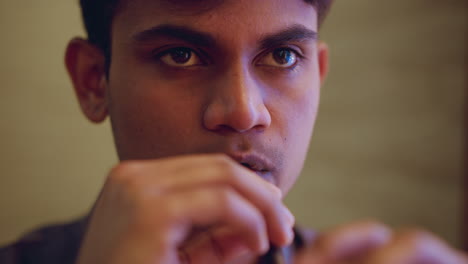 Close-up of young man holding cup near mouth with intense thoughtful eyes, appearing deep in concentration and reflection, warm ambient lighting emphasizing facial details and emotional mood in quiet indoor space