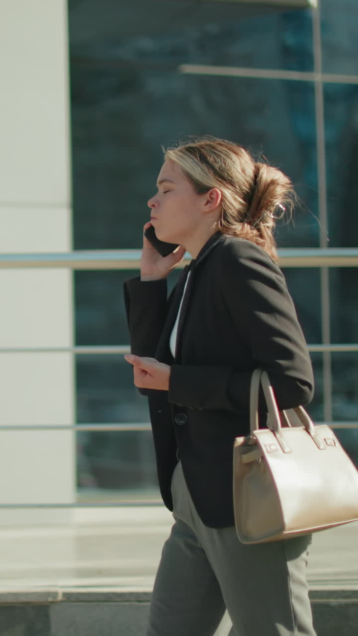 Side view of young banker on phone with customer visibly frustrated, raising hands in anger while walking past mall with modern glass facade reflecting street and urban background