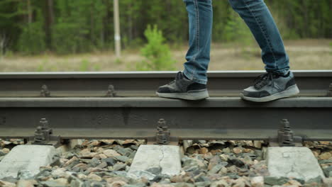 a close-up view of a man's legs in blue jeans and black sneakers walking along railway tracks, the shot focuses on the feet