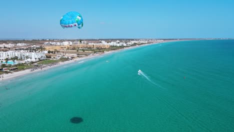 the image is an aerial view of a beach with crystal blue water of a hotel at mahdia, where people are enjoying various activities. a few surfboards can also be seen floating on the water.